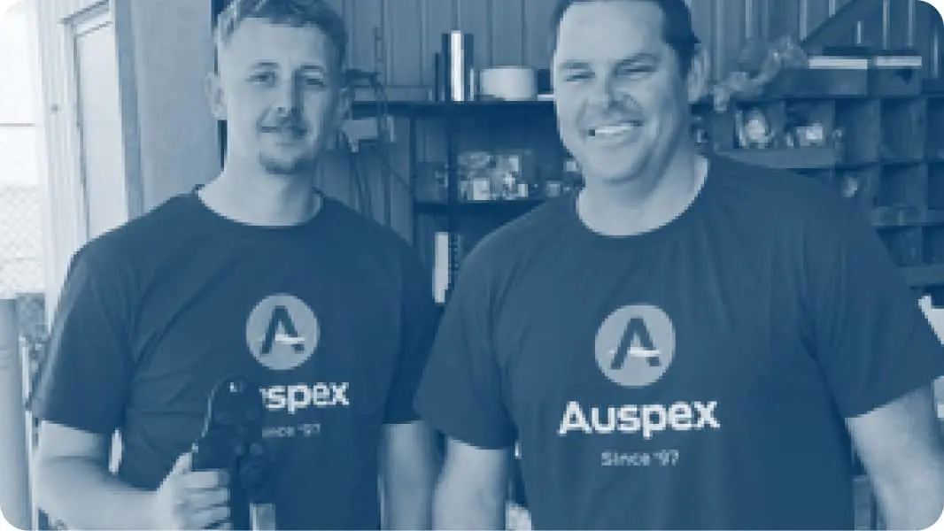 Two people standing side by side wearing matching dark Auspex t-shirts with logo, in what appears to be a workshop.