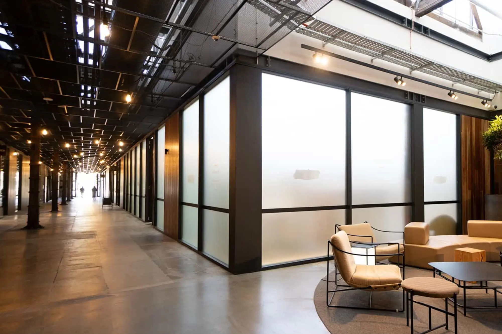 Indoor space with a long corridor, frosted glass partitions, concrete floors, and a seating area with tan chairs and small tables.