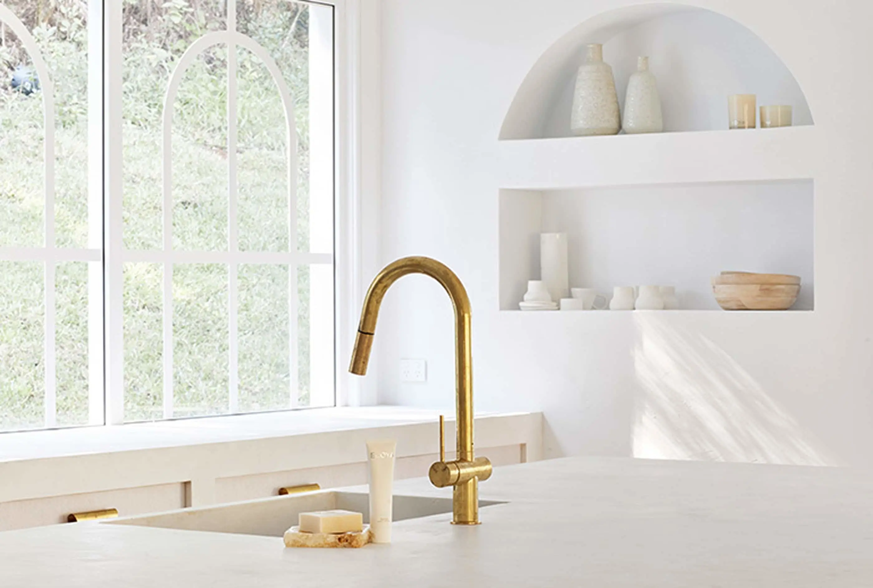 Minimalist white kitchen with brass faucet, sink, arched window, and built-in wall shelves displaying neutral decor items.