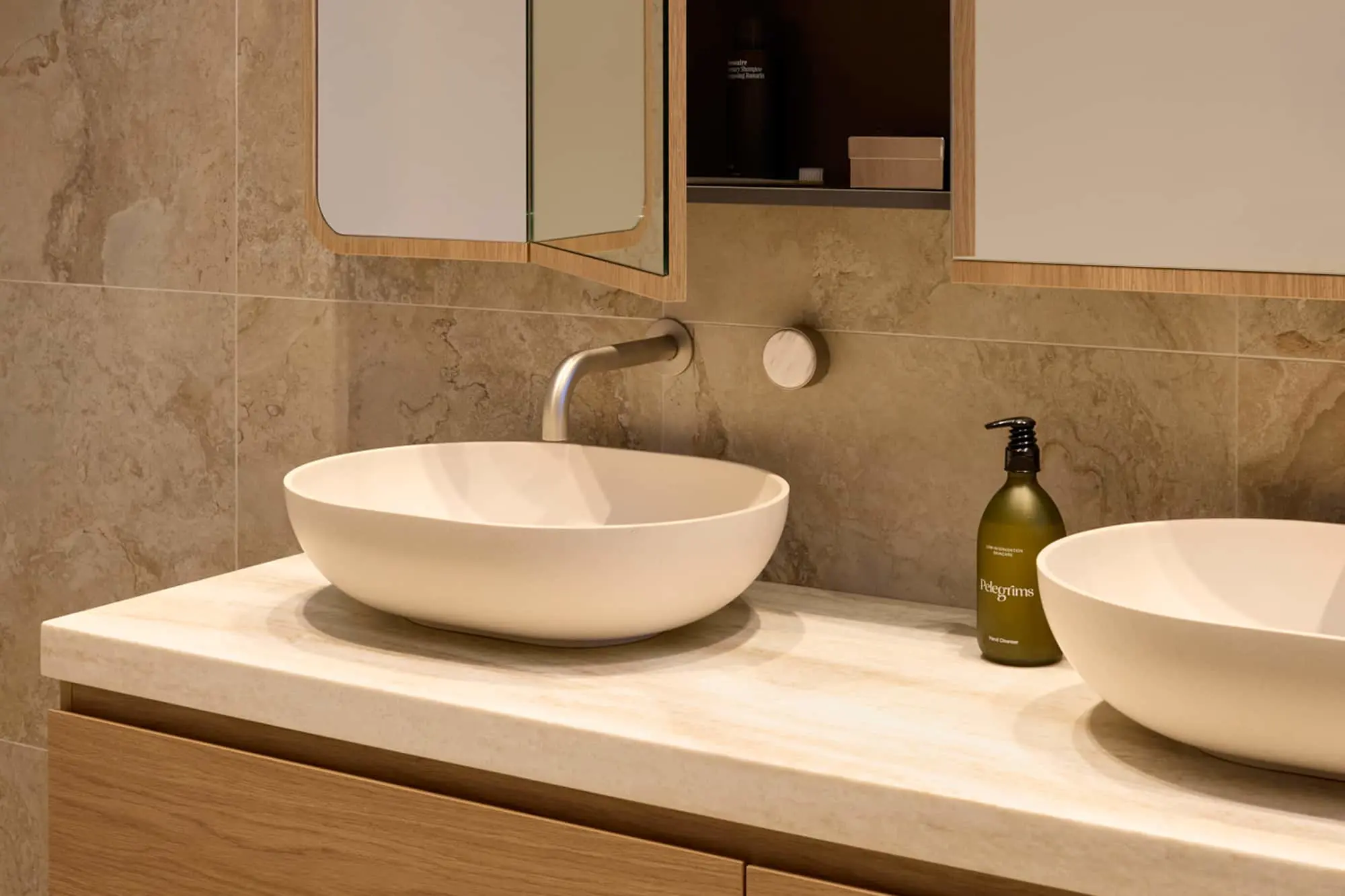 Modern bathroom with white vessel sink, wall-mounted faucet, beige stone tiles, and wooden vanity with mirror cabinet.