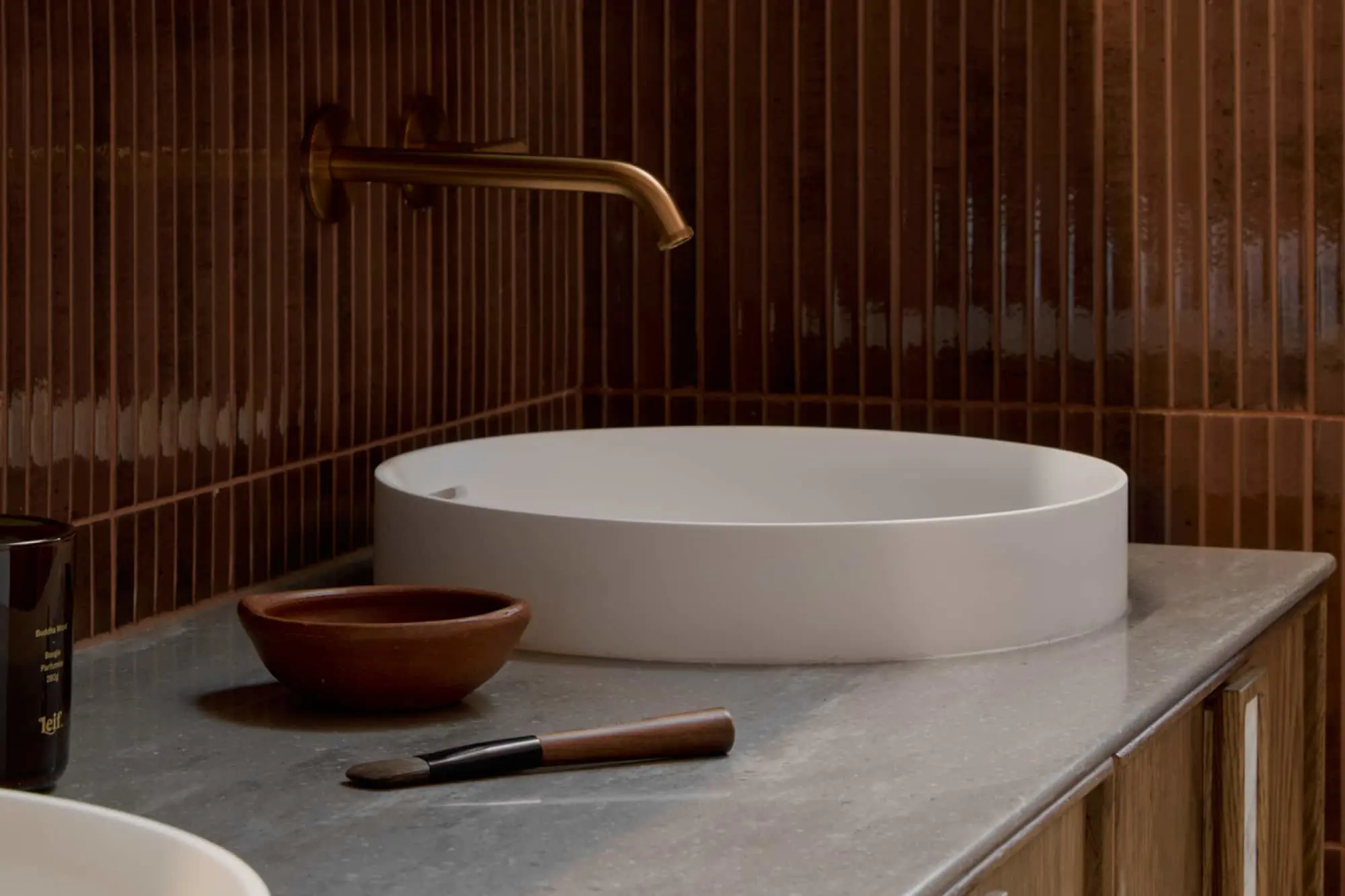 Modern bathroom with white vessel sink, brass wall-mounted faucet, wooden bowl, and brush on marble countertop against brown tile.