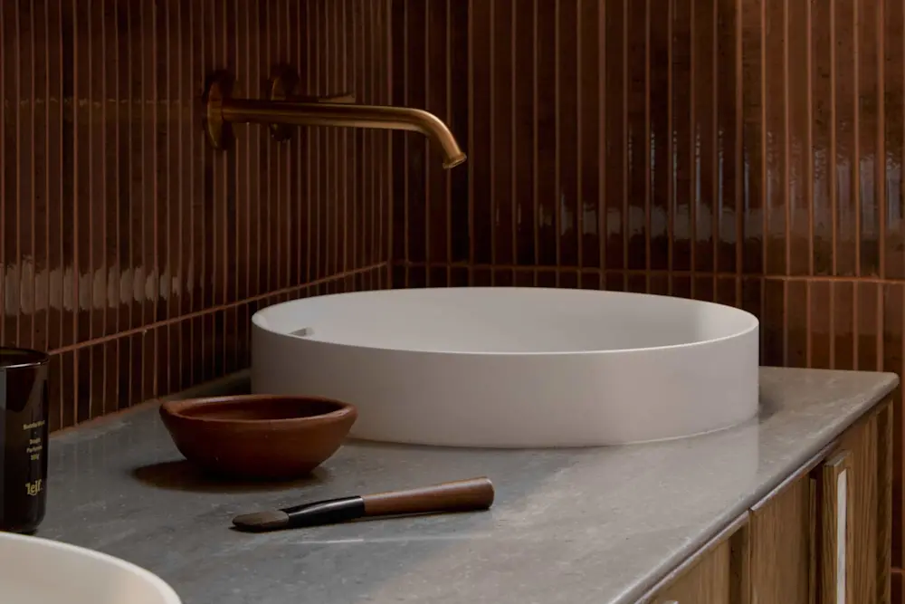 Modern bathroom with white vessel sink, brass wall-mounted faucet, wooden bowl, and brush on marble countertop against brown tile.