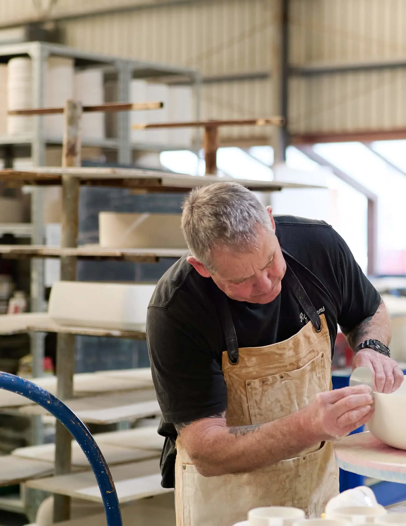 Person in tan apron working with pottery in a workshop with shelving units in the background.