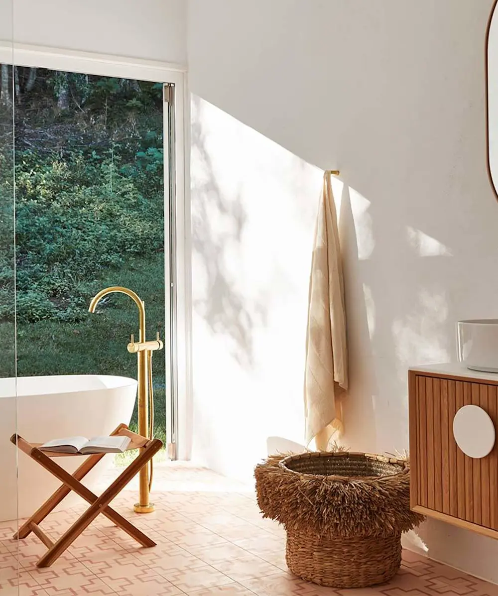 Modern bathroom with white tub, gold fixtures, wooden stool with book, and large window overlooking green forest.