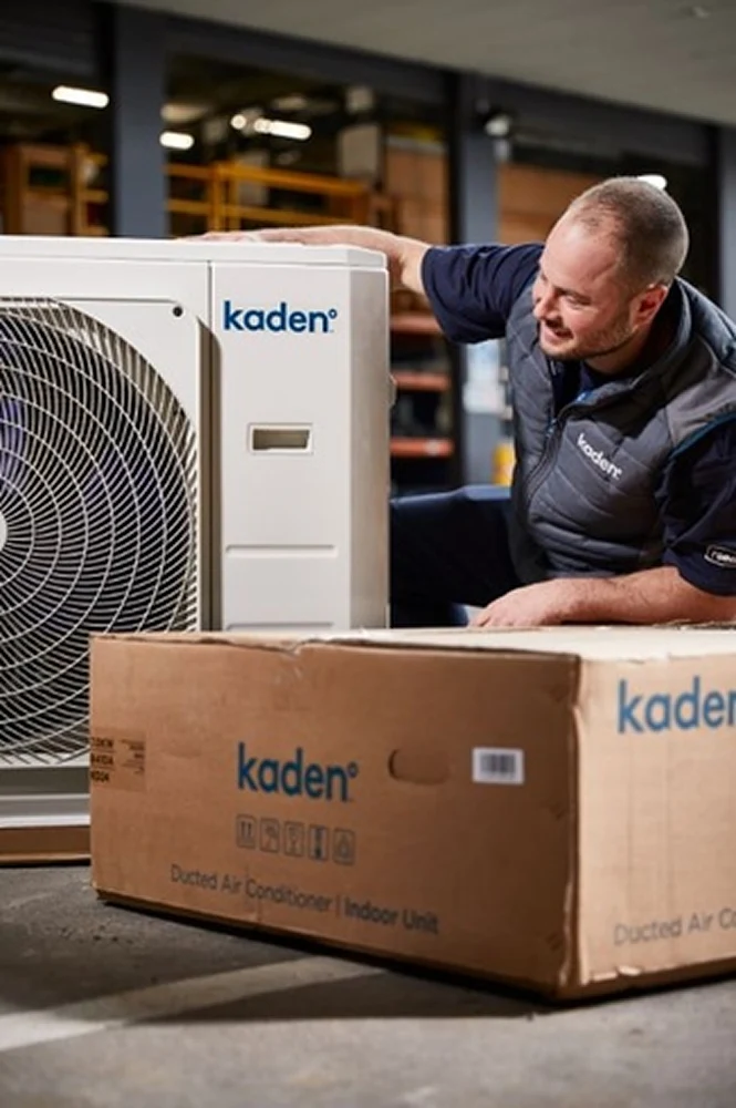 Technician inspecting a Kaden air conditioning outdoor unit with packaging box for indoor unit in a warehouse setting.