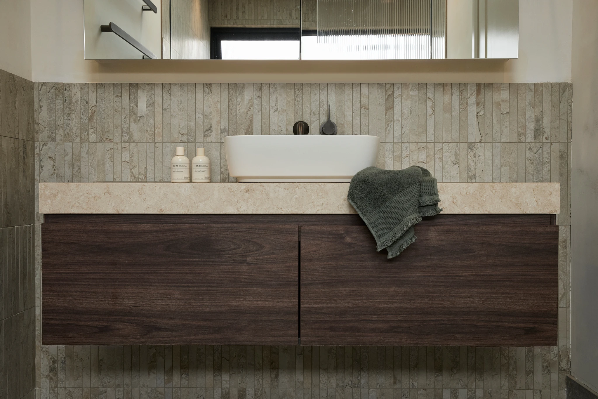 Modern bathroom vanity with white vessel sink, dark wood cabinets, stone countertop, and green towel on beige tiled wall.