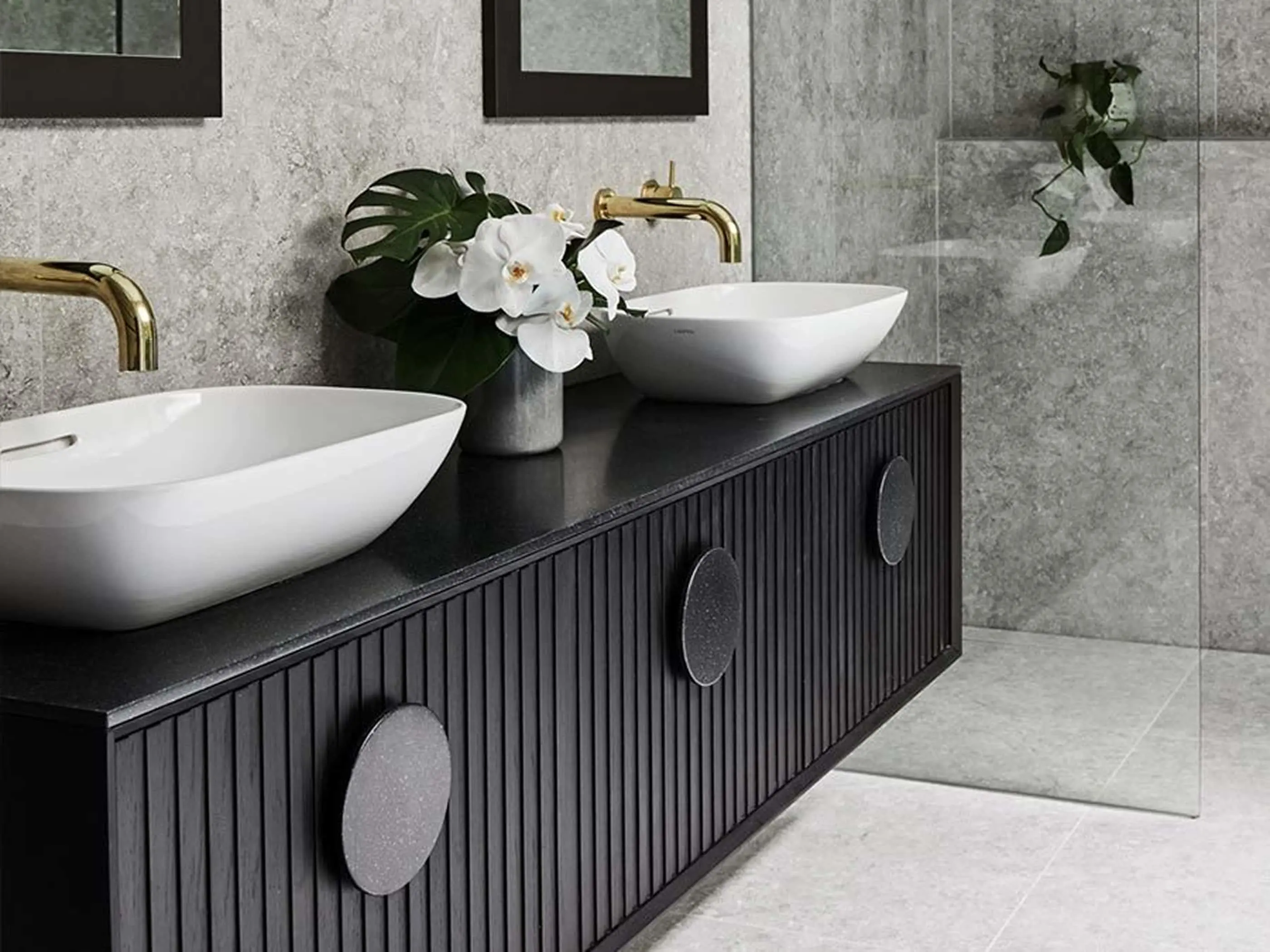 Modern bathroom with black vanity, white vessel sinks, gold faucets, and white orchids against gray stone walls.