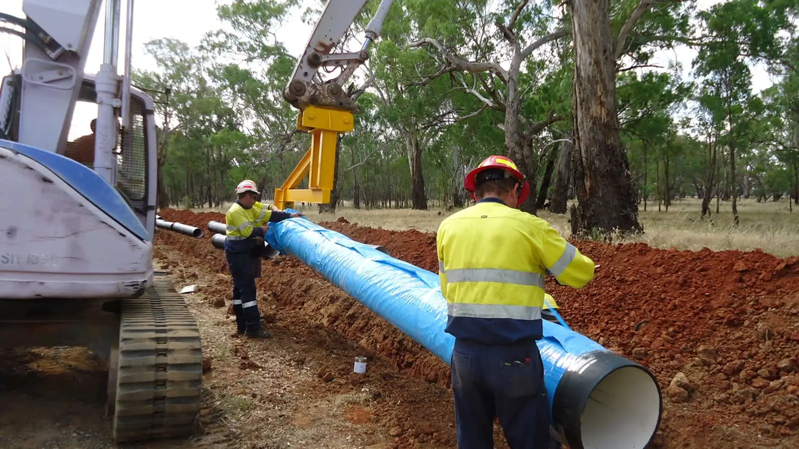 Workers in high-visibility clothing installing a large blue pipeline in a trench with excavation equipment in wooded area.