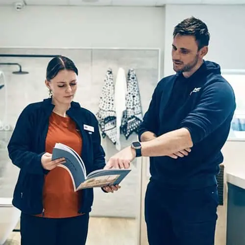 Two people reviewing a catalog together in a modern bathroom showroom with white tile walls and hanging towels.