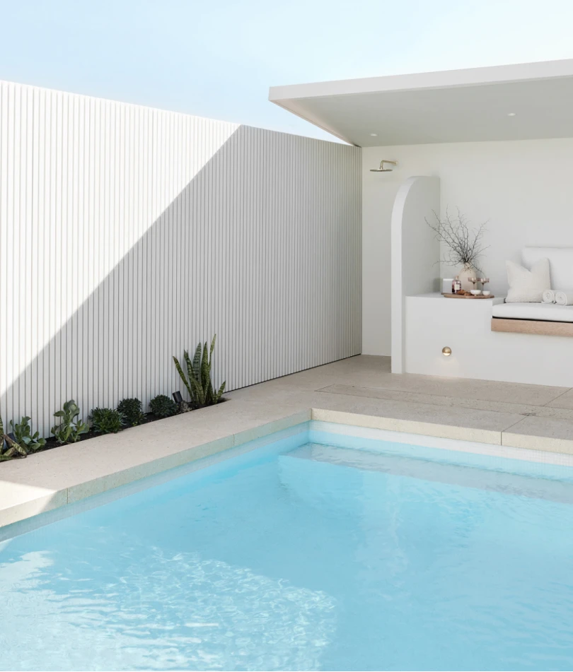 Modern minimalist pool area with white walls, blue water, and a cozy seating nook with small plants along the edge.