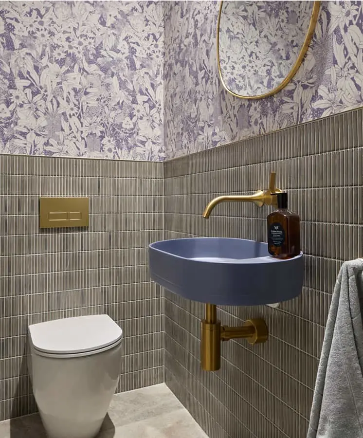 Modern bathroom with purple floral wallpaper, gray tile walls, blue sink with gold faucet, white toilet, and round mirror.