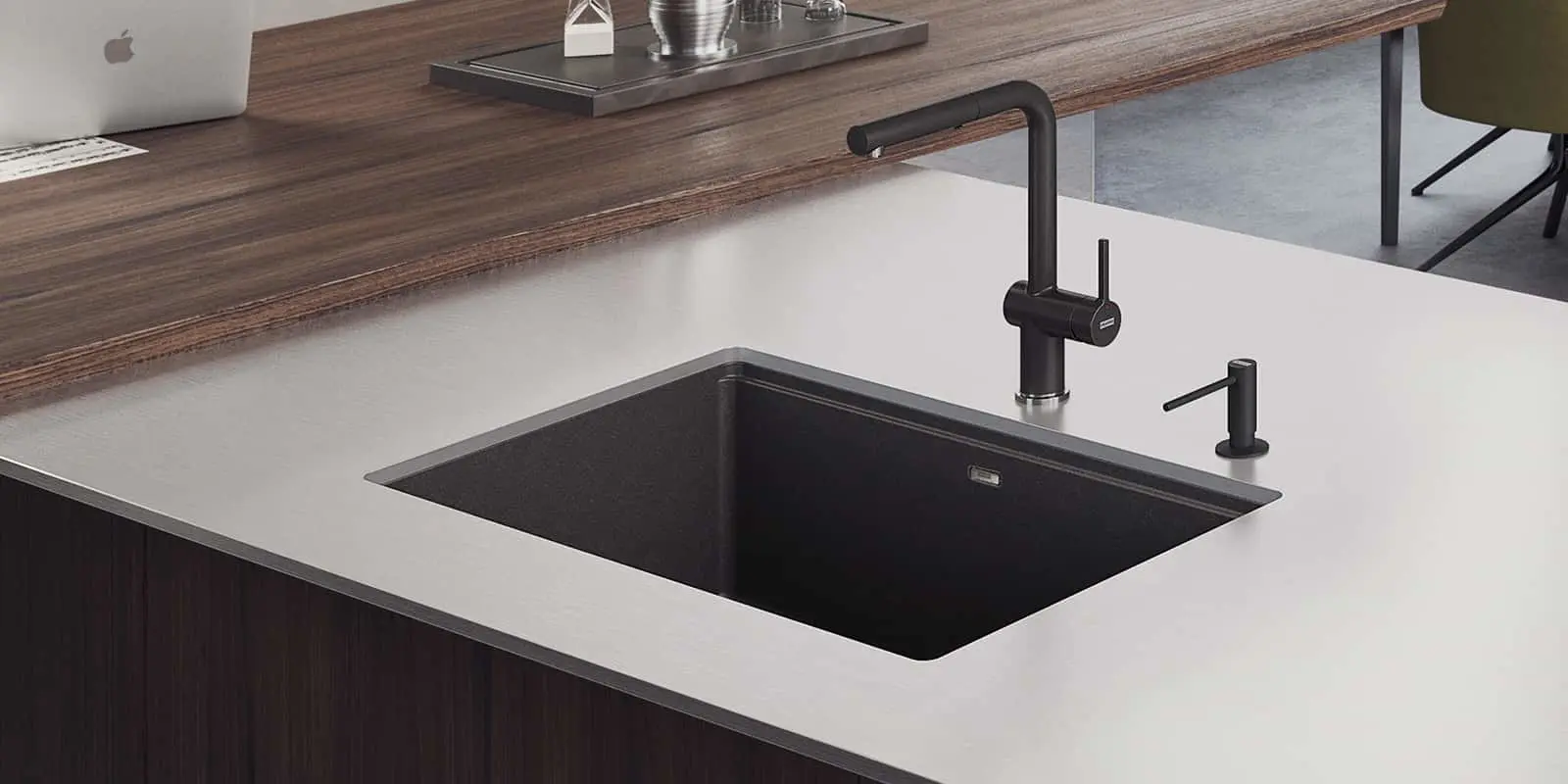 Modern kitchen with black sink and faucet in white countertop, wooden breakfast bar with laptop visible.