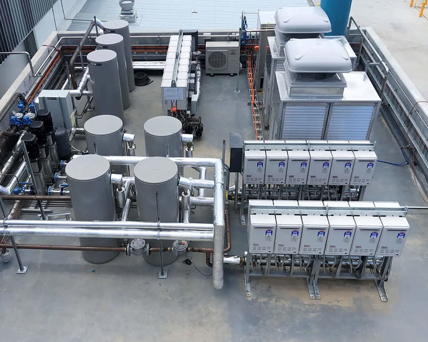 Rooftop hot water system with gray cylindrical tanks, metal pipes, control panels, and air conditioning units on concrete surface.