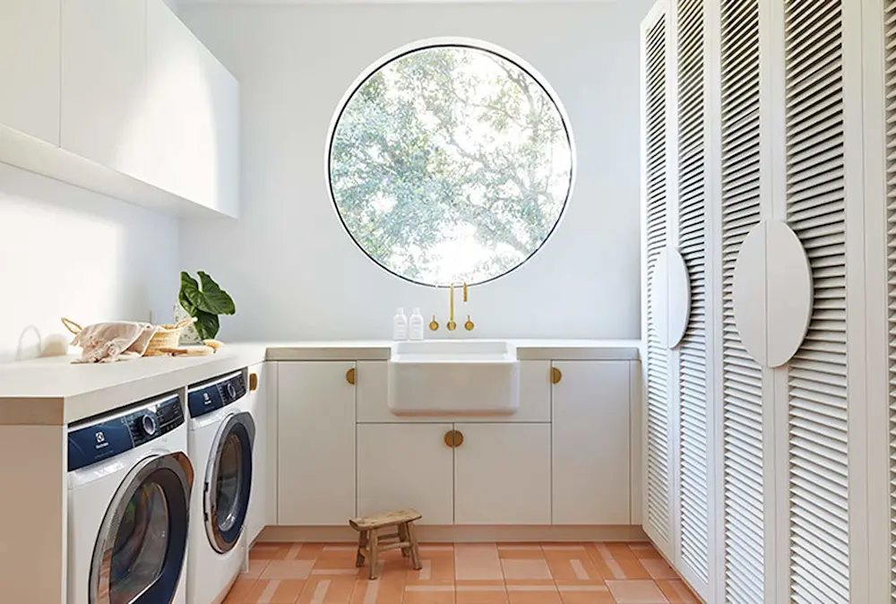 Modern white laundry room with circular window, farmhouse sink, washer and dryer, and terracotta tile floor.