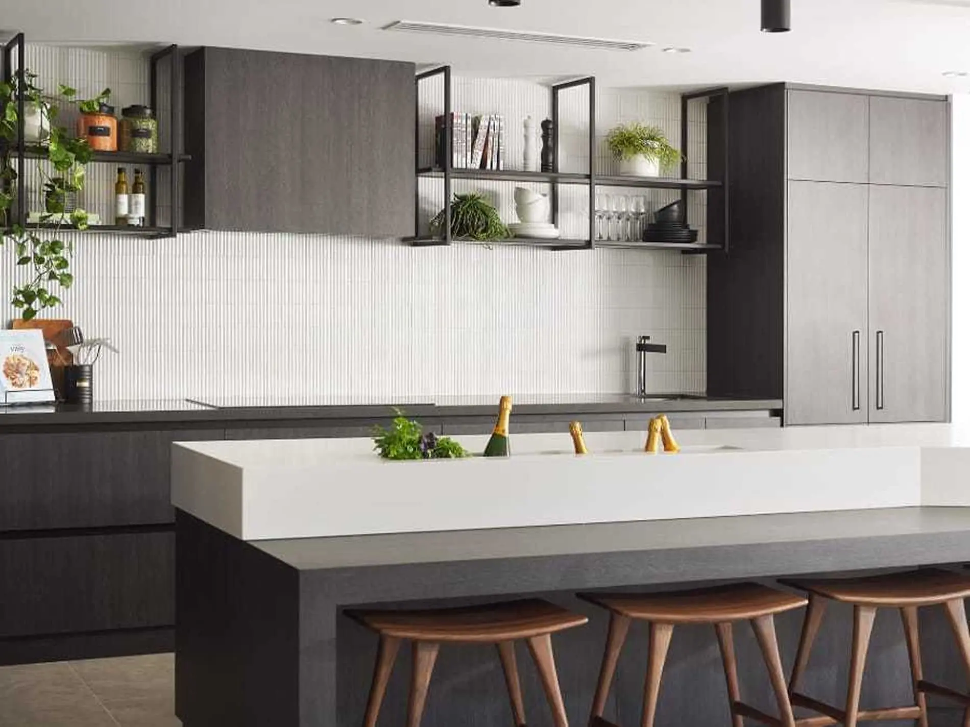Modern kitchen with gray and white cabinetry, open shelving with plants, and island with champagne bottles and wooden stools.