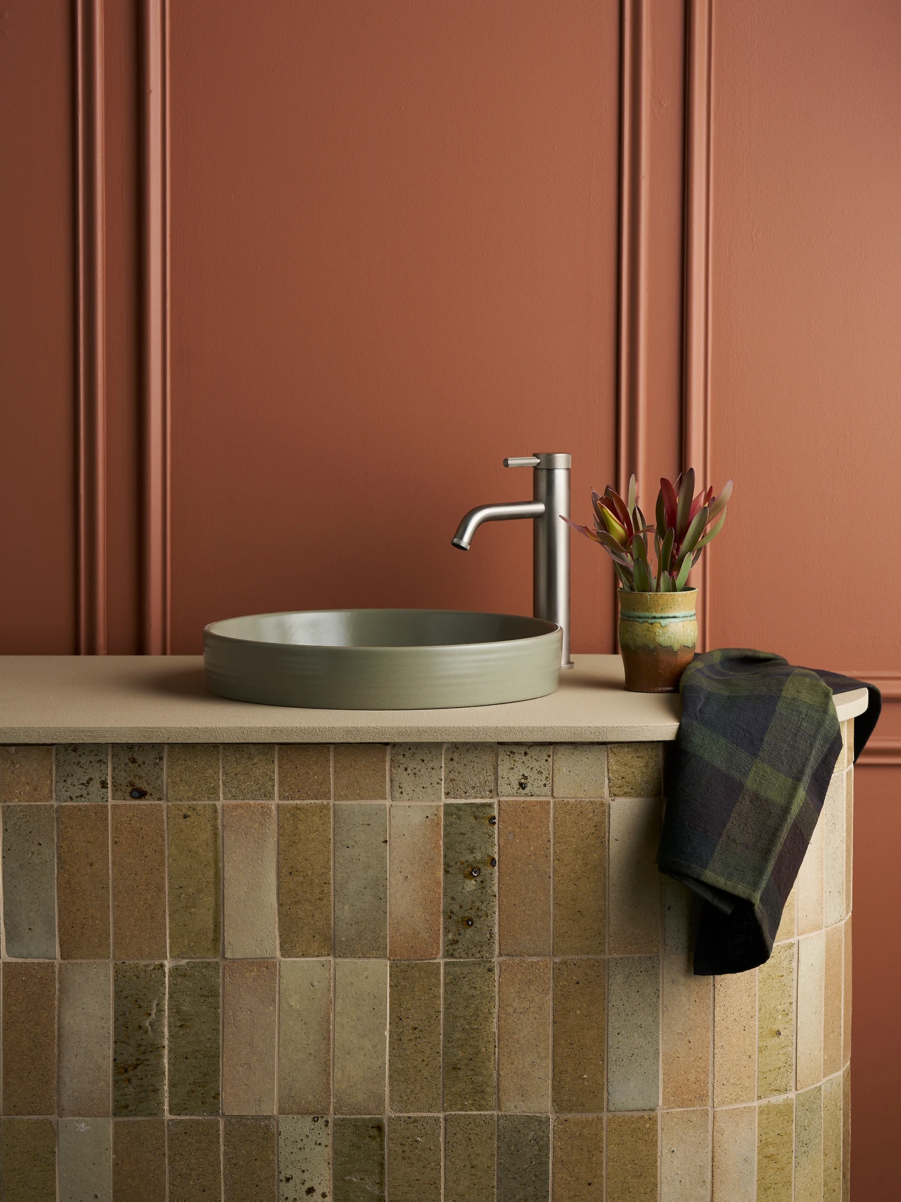 Modern bathroom with sage semi inset basin, chrome faucet, and dark towel against terracotta walls and beige tile.