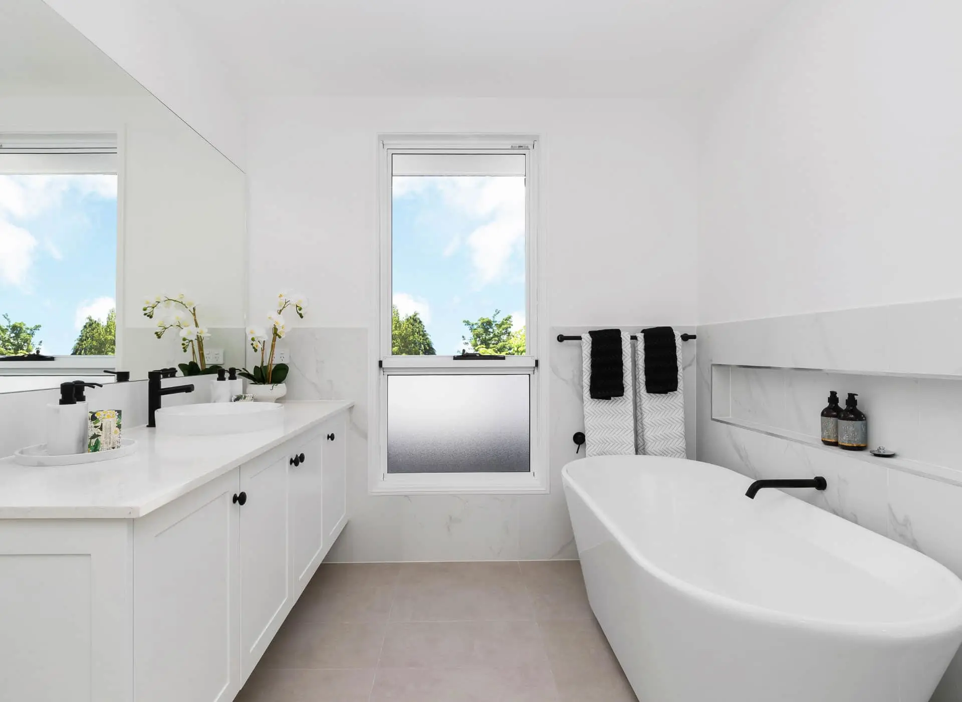 Modern white bathroom with freestanding tub, double vanity, black fixtures, and window showing blue sky and trees.