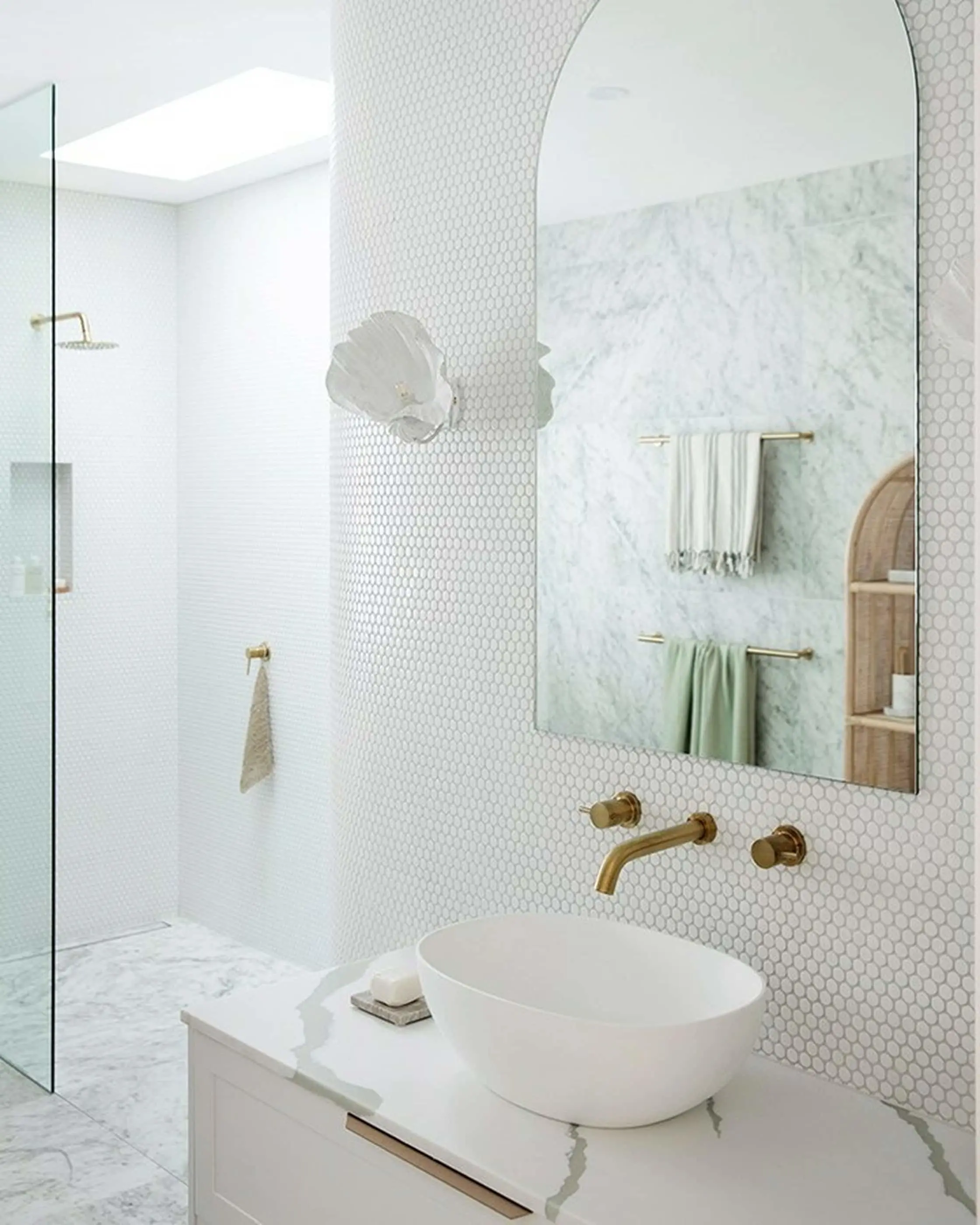 Modern white bathroom with hexagon tile walls, vessel sink, brass fixtures, arched mirror, and walk-in shower.