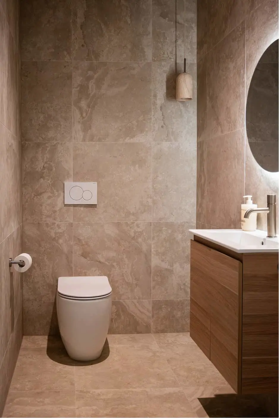 Modern bathroom with beige stone tile walls, white toilet, wooden vanity with white sink, and pendant light.