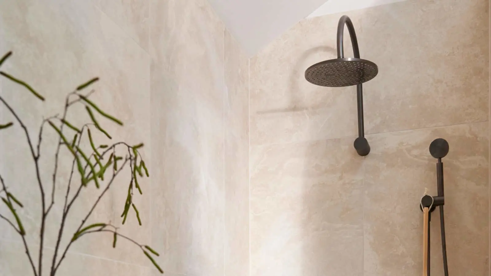 Modern shower with black rainfall showerhead and handheld attachment on beige tiled walls, green plant visible.