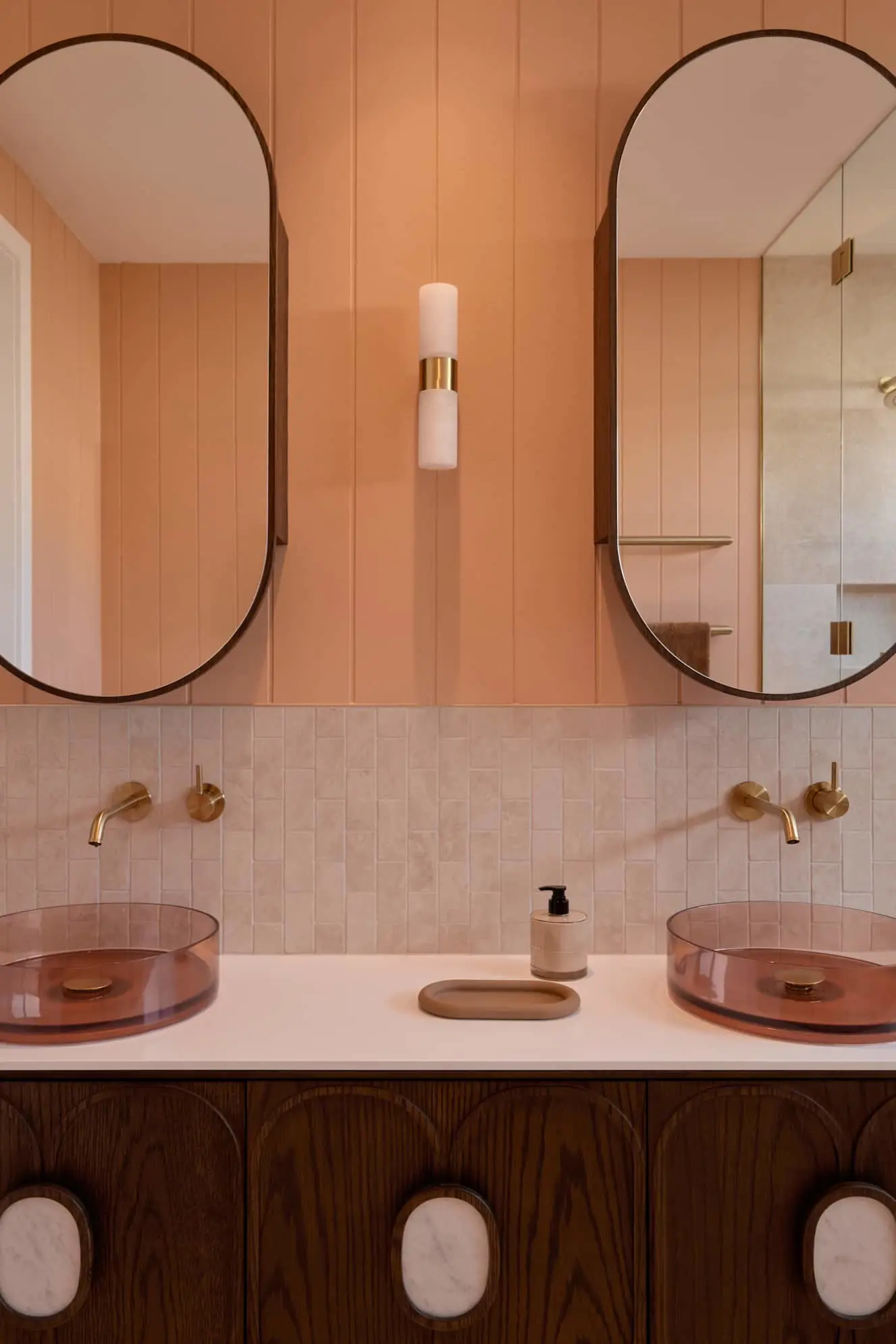 Modern bathroom with peach walls, oval mirrors, amber glass sinks, brass fixtures, and wooden cabinetry.