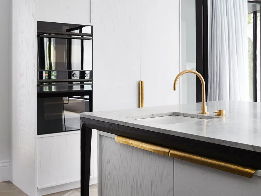 Modern kitchen with marble countertop, brass faucet and handles, built-in black ovens, and white cabinetry.