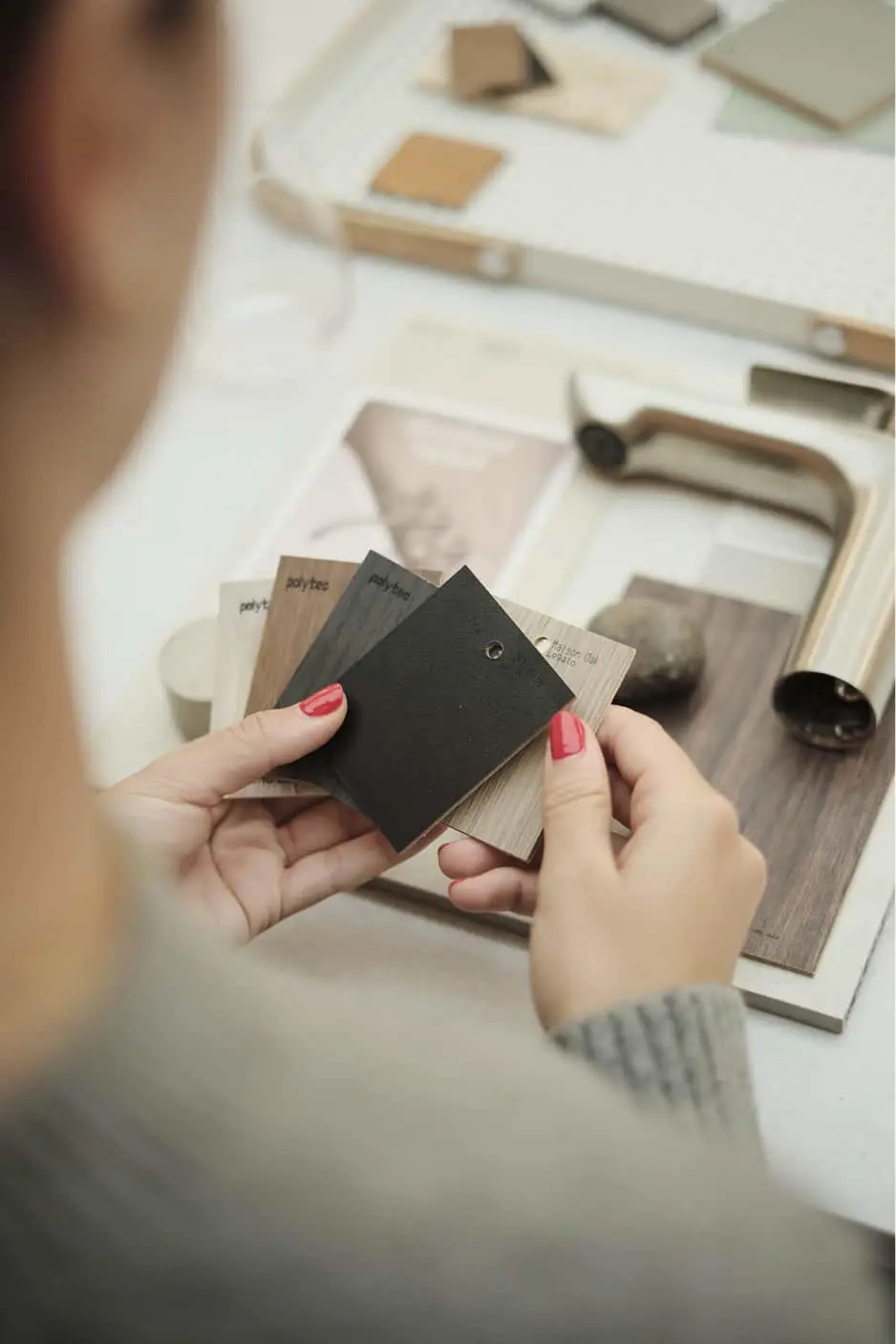 Hands with red nails holding material samples including black and wood finishes on a white workspace with design materials.