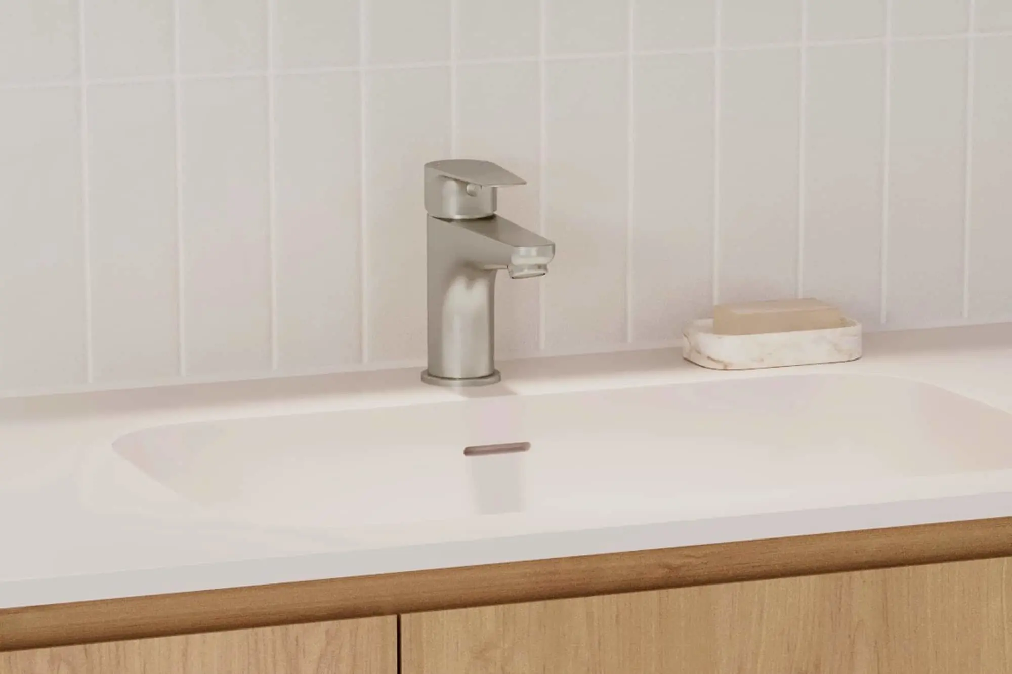 Modern brushed nickel bathroom faucet on white sink with soap dish against tiled wall and wooden cabinet below.