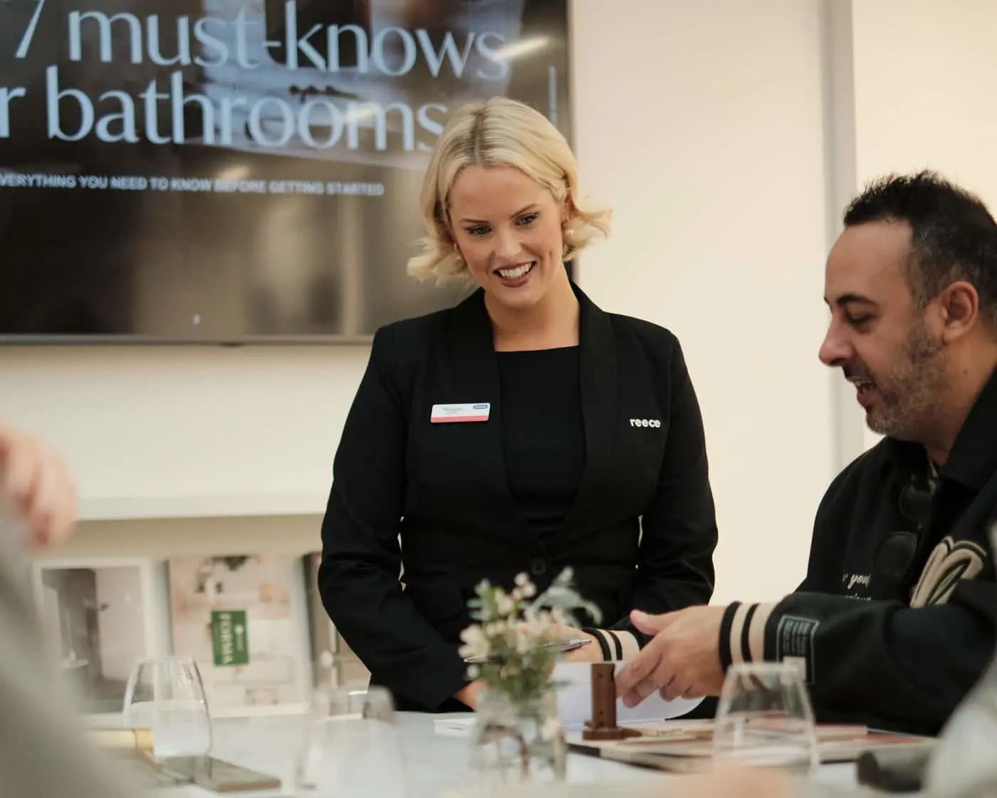 A Reece staff member in a black blazer smiles while speaking with a seated customer at a table in a showroom.