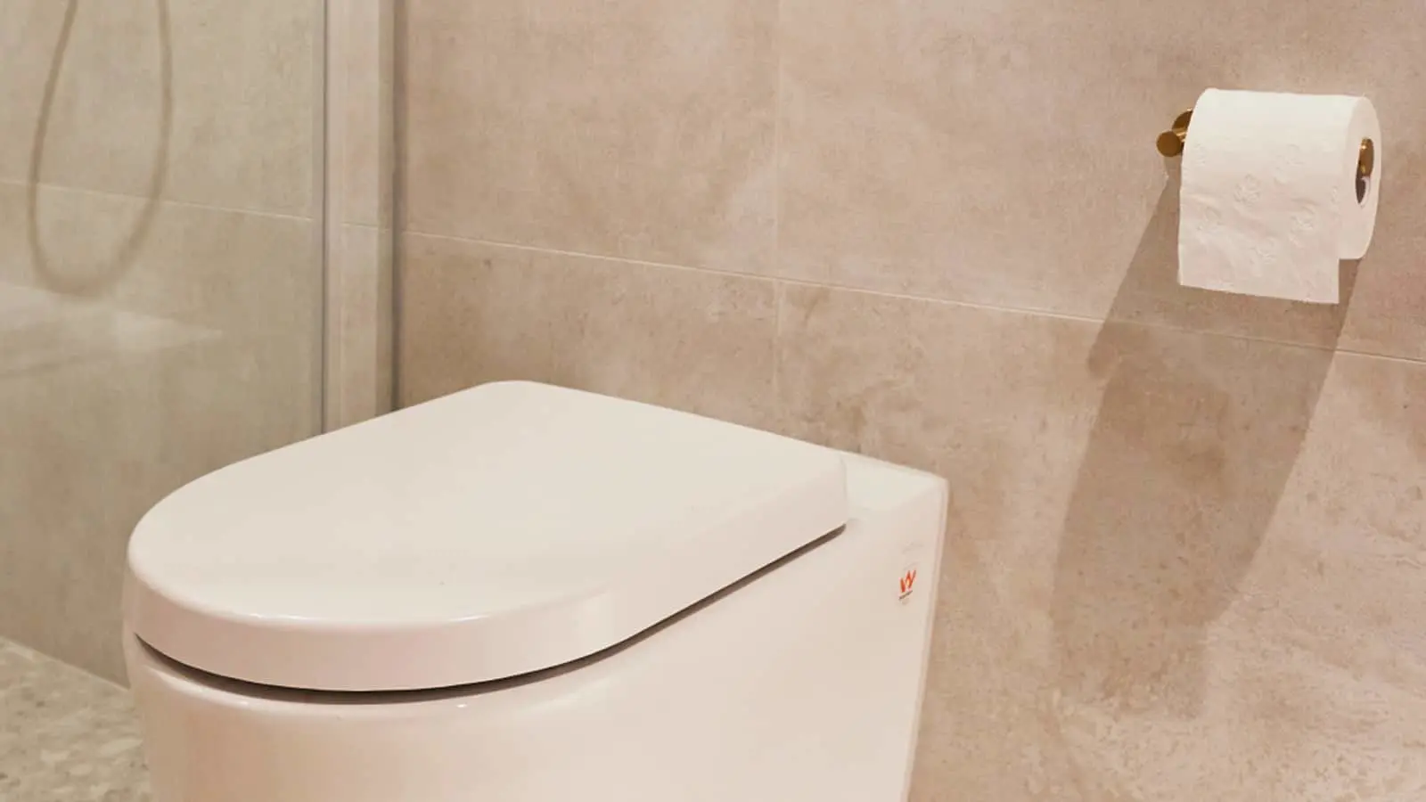 Modern white toilet with closed lid in a bathroom with beige tiled walls and toilet paper holder mounted nearby.