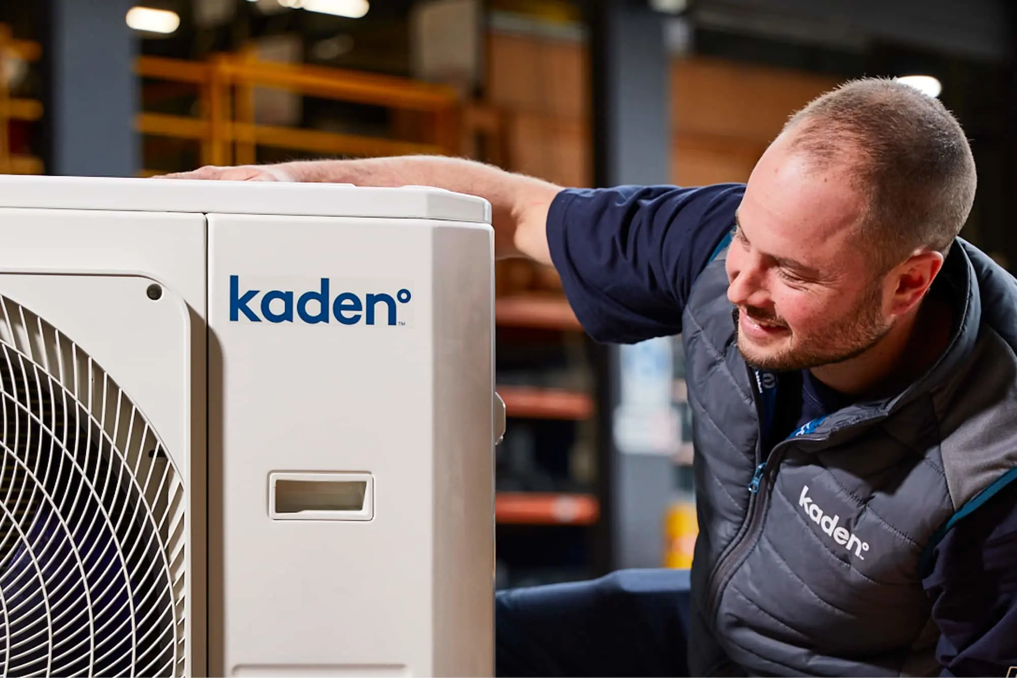 A man in a Kaden vest inspects a white Kaden branded outdoor heat pump unit in a warehouse setting.