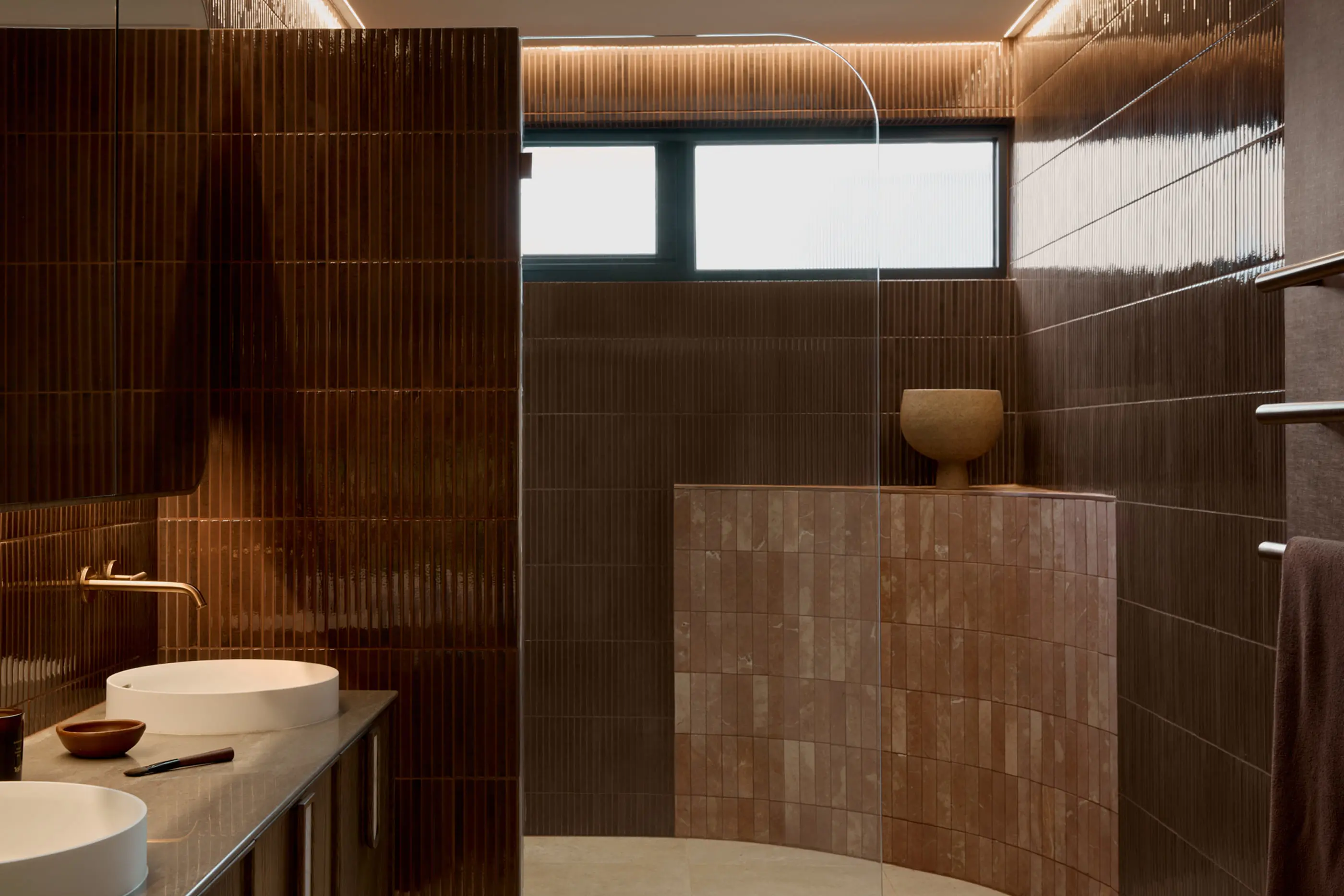 Modern bathroom with dark brown tiled walls, white sink, curved shower partition, and natural light from window above.