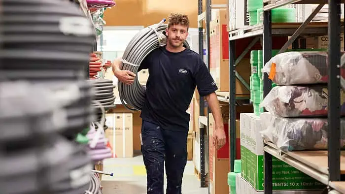 Worker in navy shirt carrying coiled pipe through warehouse aisle with shelves of construction supplies and boxes