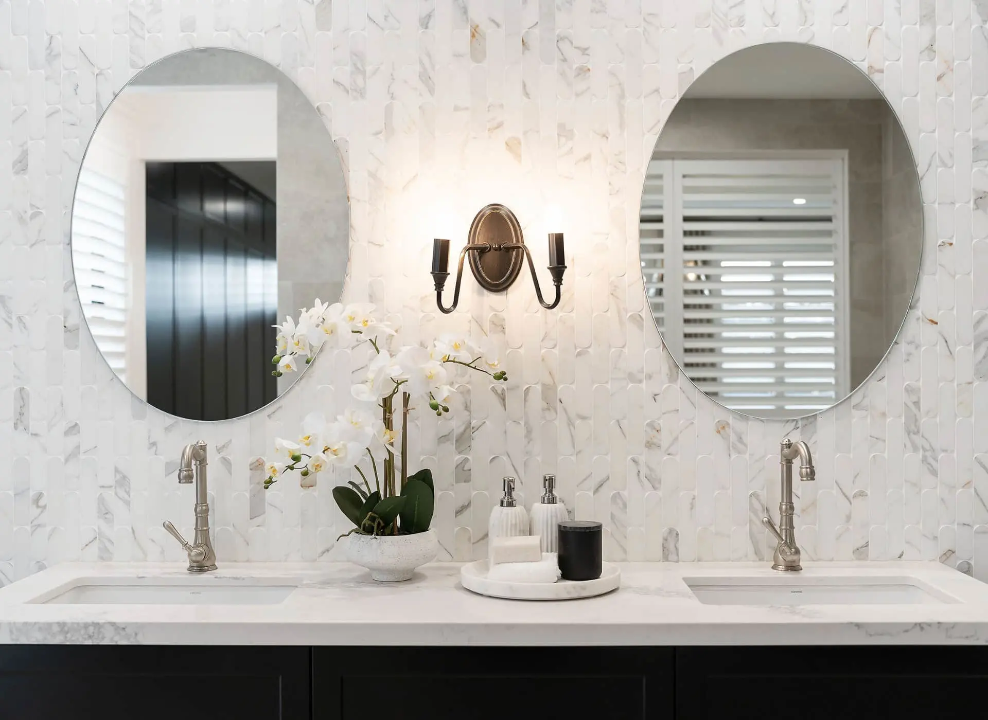 Elegant bathroom with dual round mirrors, marble tile wall, white orchid plant, and brass wall sconce between sinks.