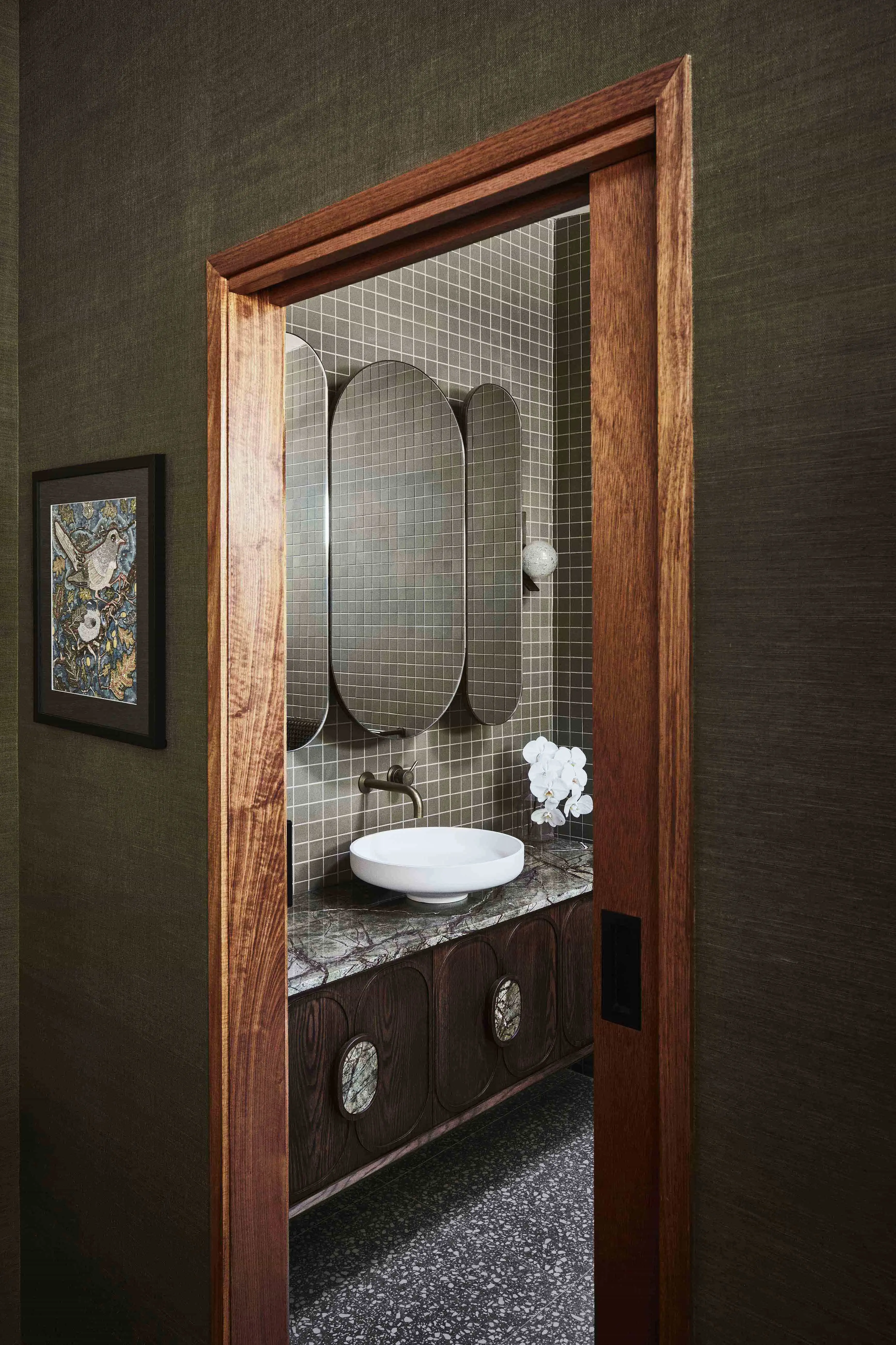 View through a wood-framed doorway into a bathroom with green tiles, oval mirrors, a white vessel sink, and a dark wood vanity.