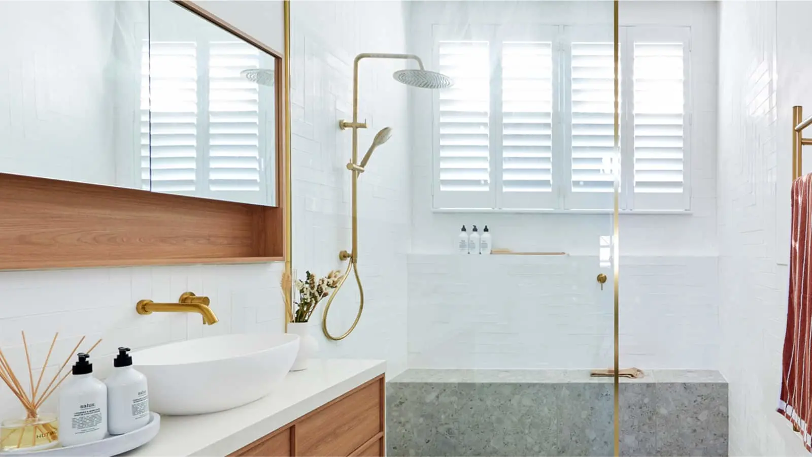 Modern white bathroom with wooden vanity, gold fixtures, glass shower enclosure, and white plantation shutters.