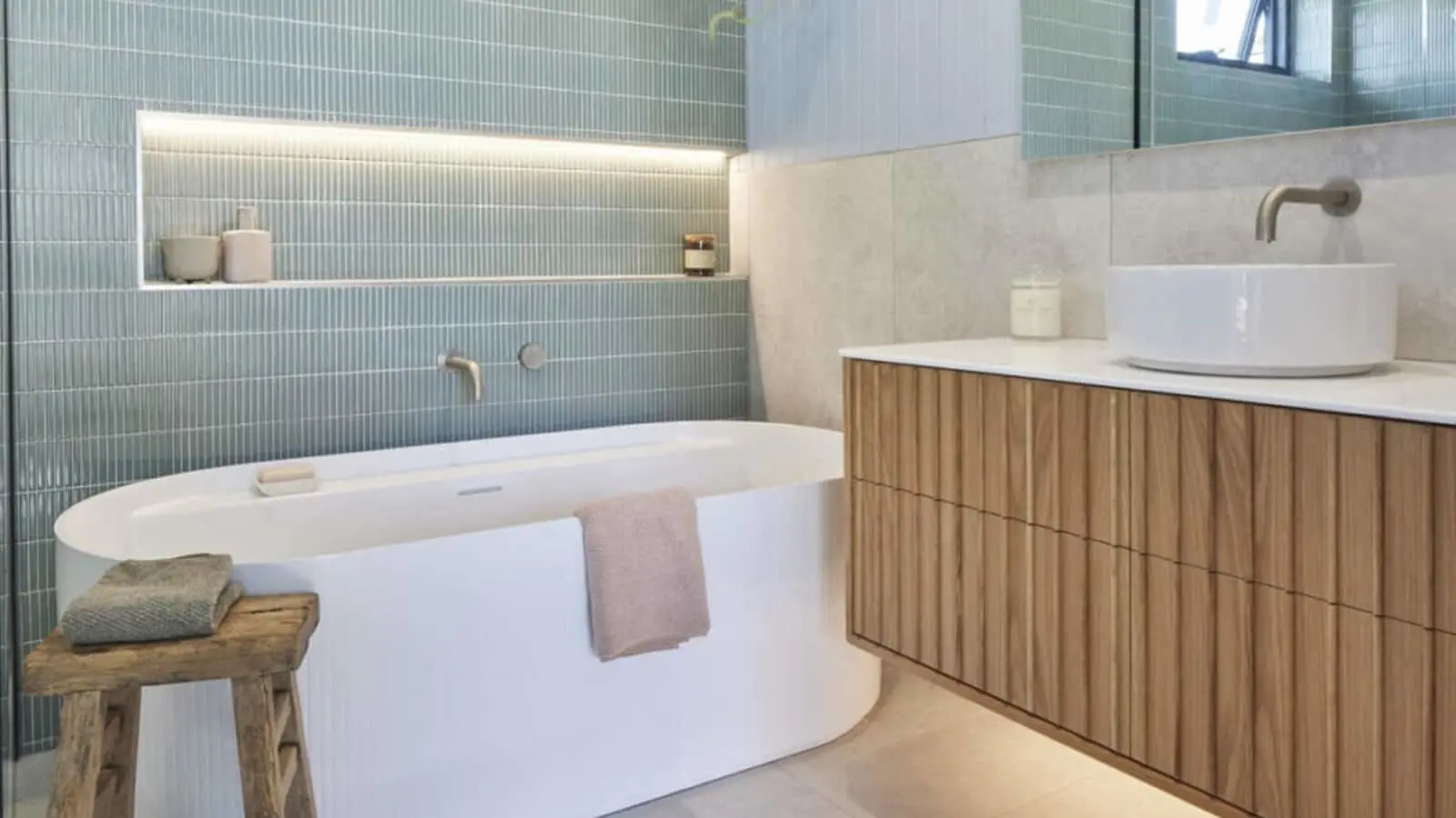 Modern bathroom with white bathtub, blue-green tiled wall, wooden vanity, and illuminated shelf niche.