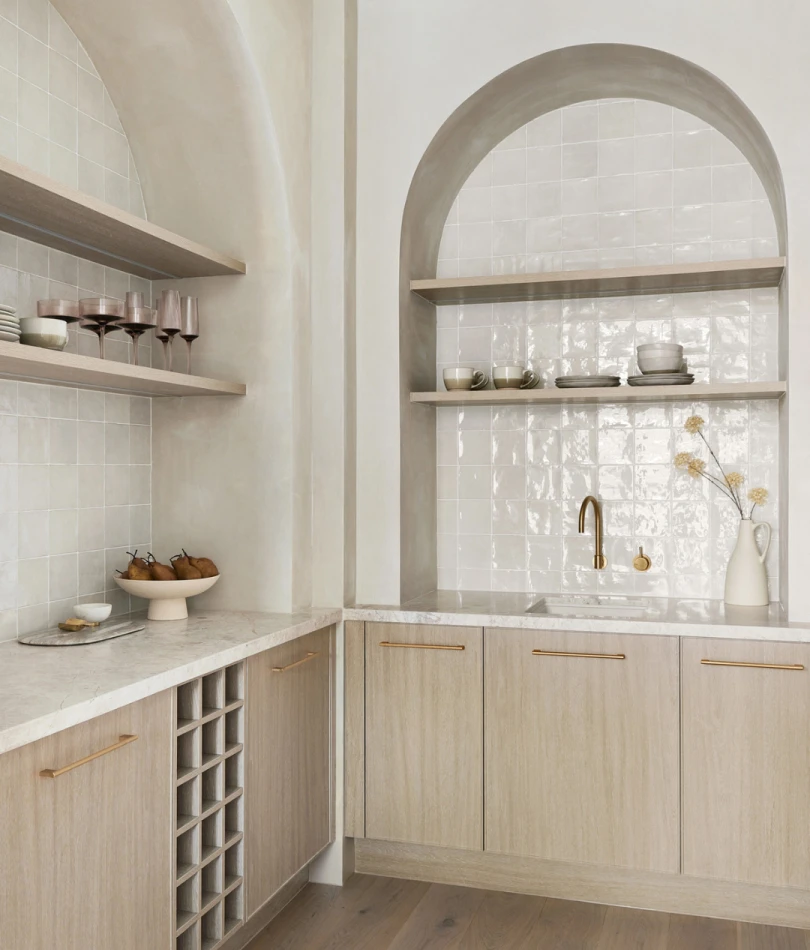 Modern kitchen with arched niche, white tile backsplash, light wood cabinets, gold fixtures, and open shelving displaying dishware.