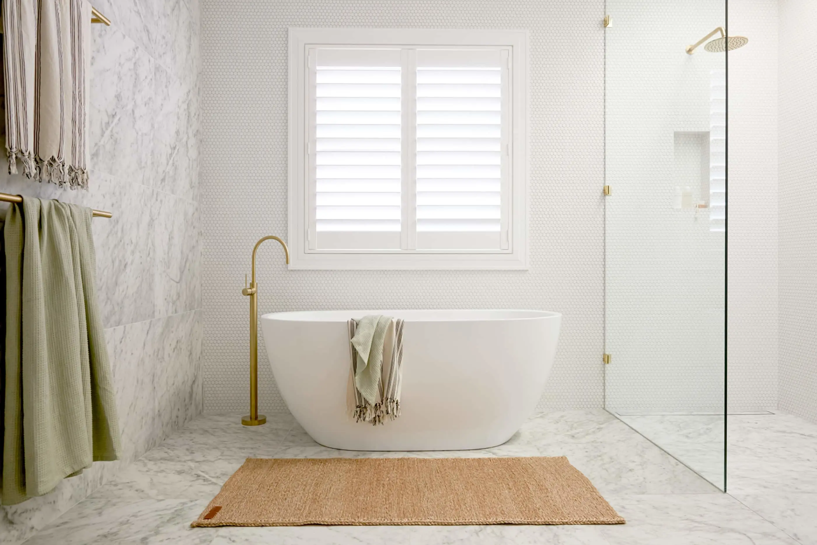 Modern white bathroom with freestanding tub, marble walls, white shuttered window, and brass fixtures.