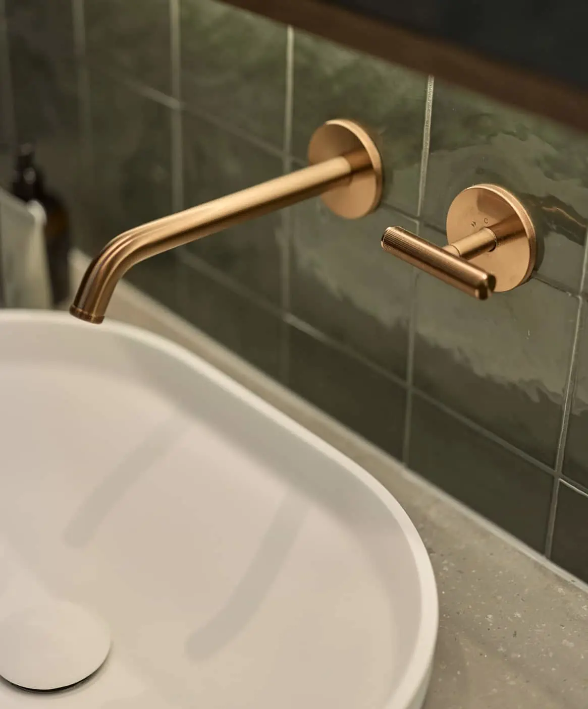 Bronze wall-mounted bathroom faucet with separate handle against green tile wall, next to white sink basin.