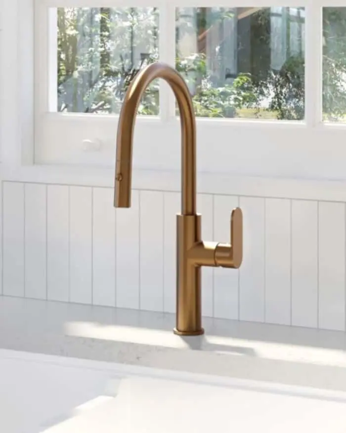 Modern brass kitchen faucet with pull-down sprayer on white countertop, window with garden view in background.