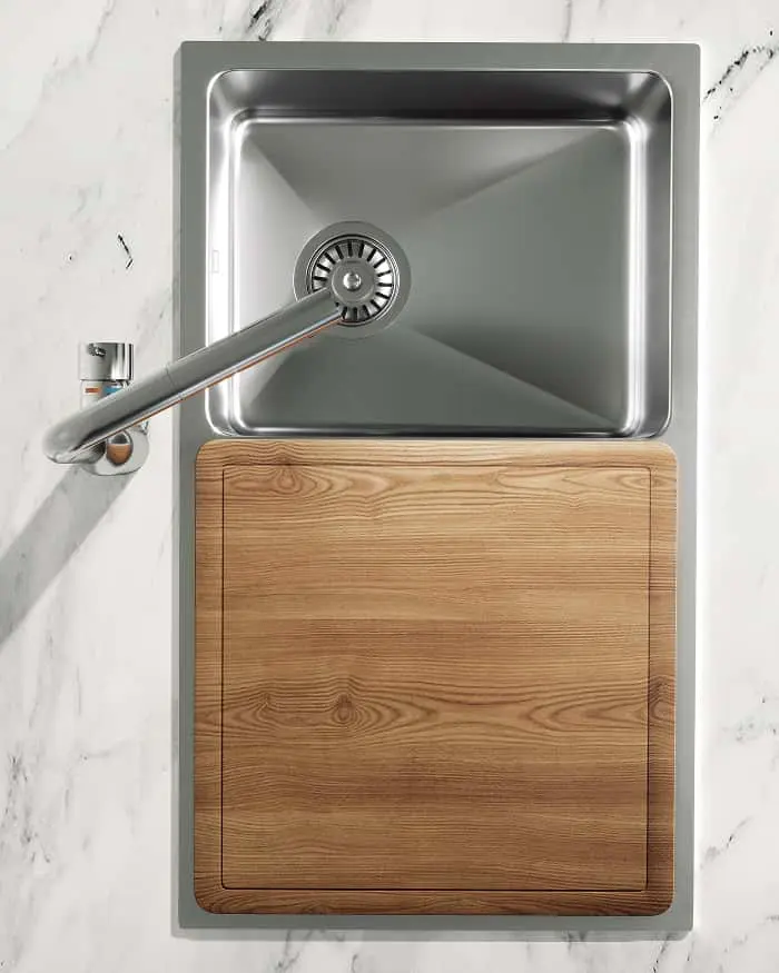 Stainless steel kitchen sink with faucet and wooden cutting board on marble countertop, viewed from above.