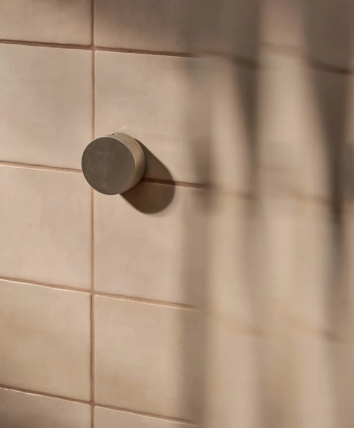 Circular shower mixer on beige tiled bathroom wall with window shadow patterns visible.