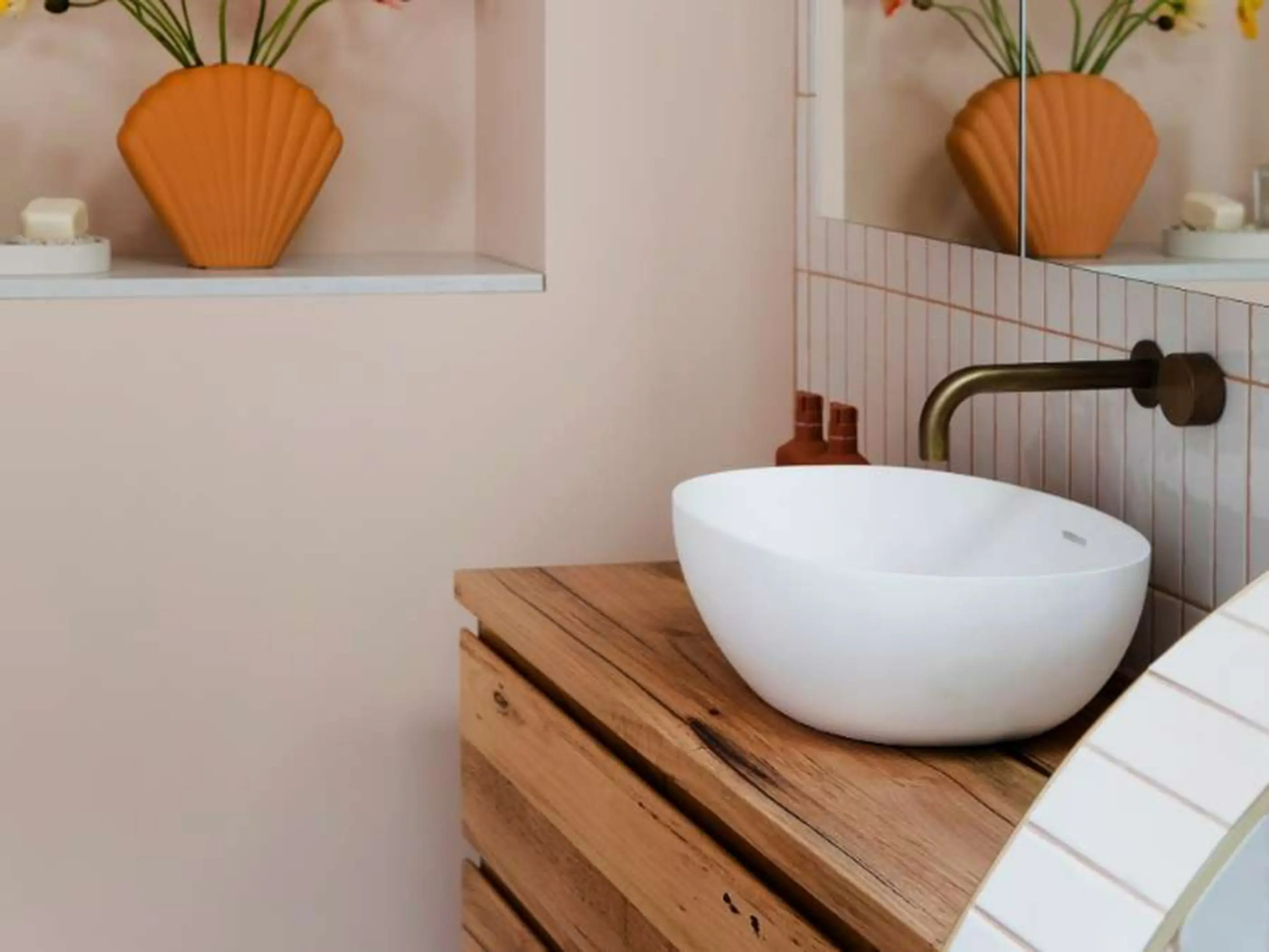 Modern bathroom with white vessel sink on wooden vanity, brass faucet, and orange shell-shaped vase with flowers on shelf.