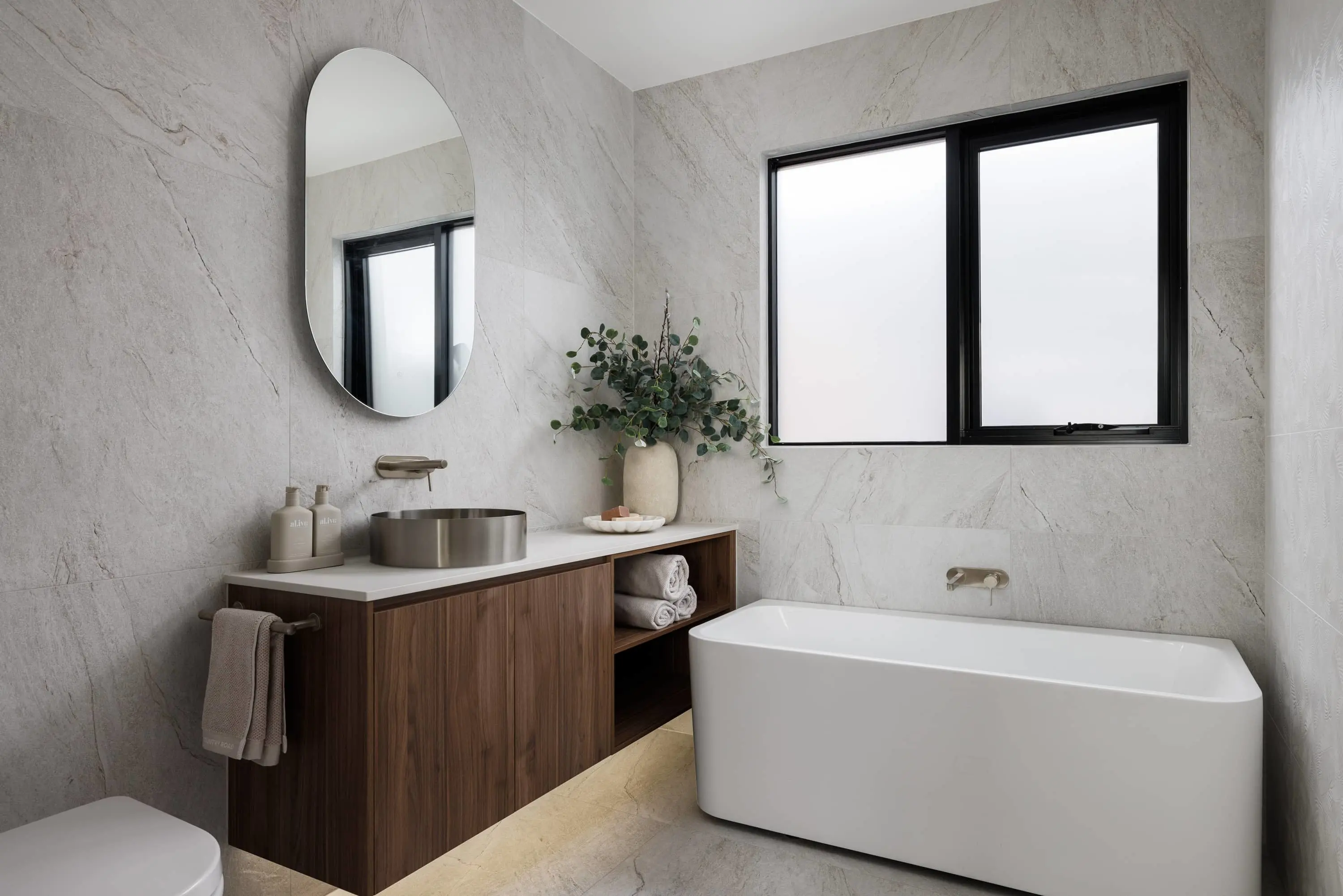 Modern bathroom with marble walls, wooden vanity, oval mirror, metal sink, white bathtub, and black-framed window.