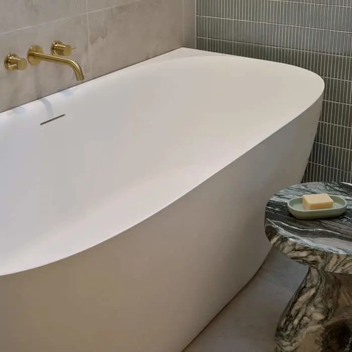 Modern white freestanding bathtub with gold wall-mounted faucet and marble side table holding soap.