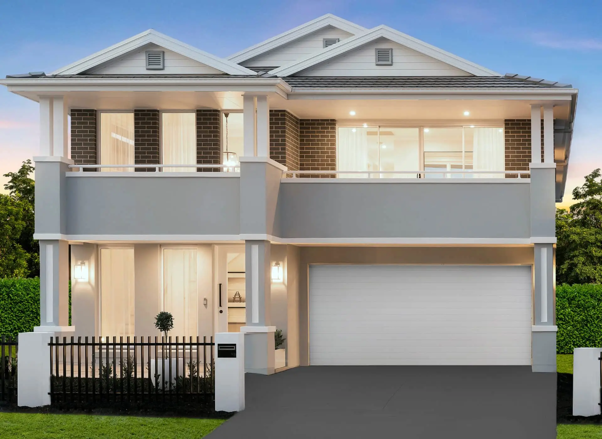 Modern two-story home with white and gray exterior, brick accents, balconies, and attached garage, illuminated at dusk.