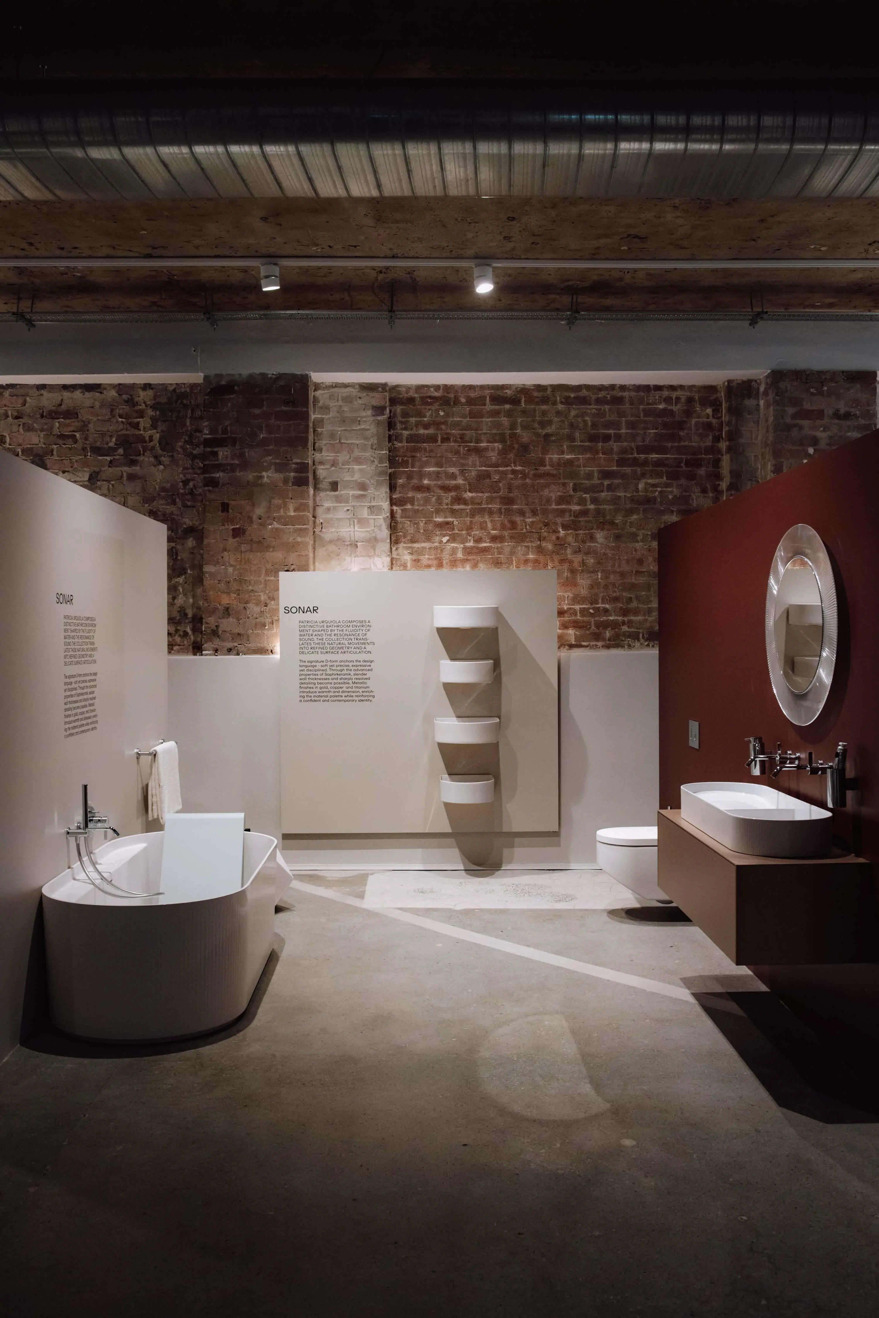 Modern bathroom showroom with white fixtures, exposed brick walls, and a red accent wall featuring SONAR collection displays.
