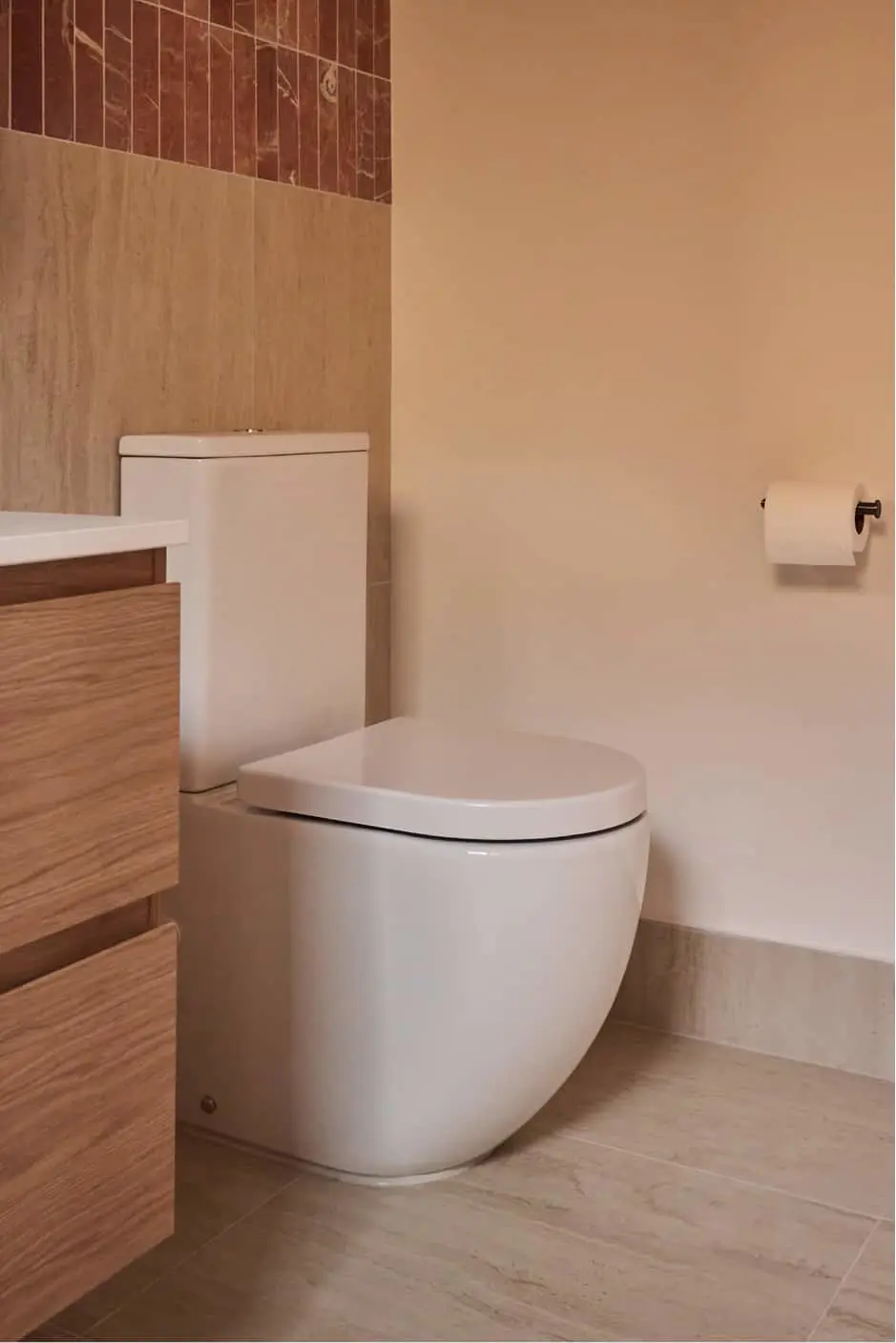 Modern bathroom with white toilet, wood vanity, beige walls, brown tile accent, and toilet paper holder.