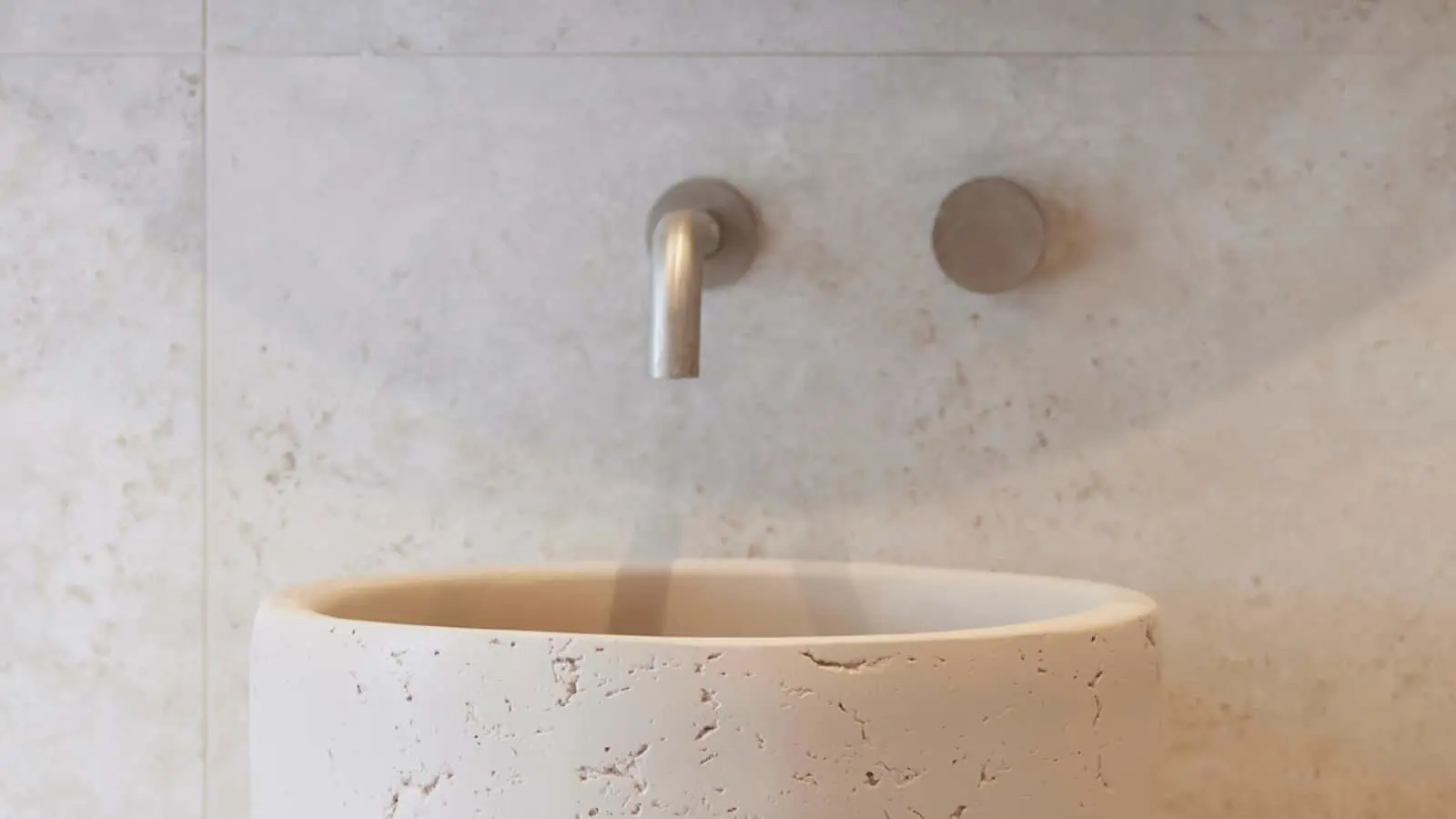 Modern stone bathroom sink with wall-mounted stainless steel faucet against light marble wall.